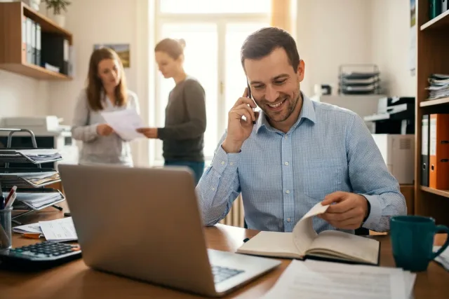 Obchodník telefonuje se zákazníkem u notebooku, v pozadí kolegyně probírá papíry s dalším klientem.