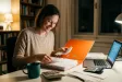 Žena u stolu třídí faktury a účtenky do šanonu vedle notebooku a kalkulačky, usmívá se s úlevou.
