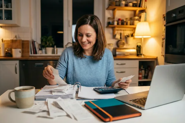 Žena u kuchyňského stolu večer třídí účtenky do šanonu vedle notebooku a kalkulačky.