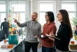 Tři kolegové v coworkingu se smějí nad plánem automatizace u skleněné stěny s poznámkami.
