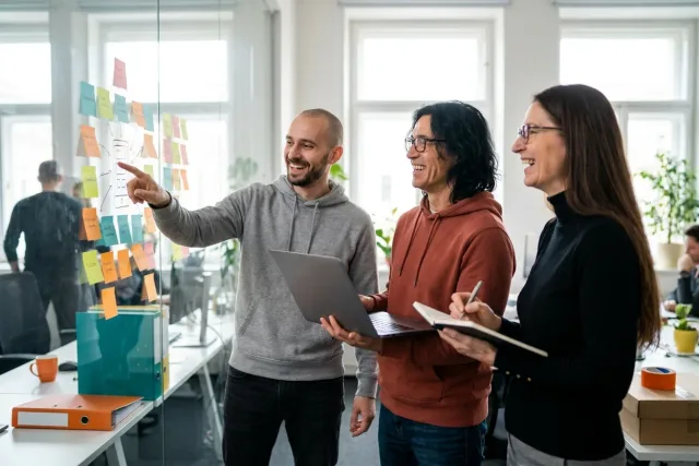 Tři kolegové v coworkingu se smějí nad plánem automatizace u skleněné stěny s poznámkami.