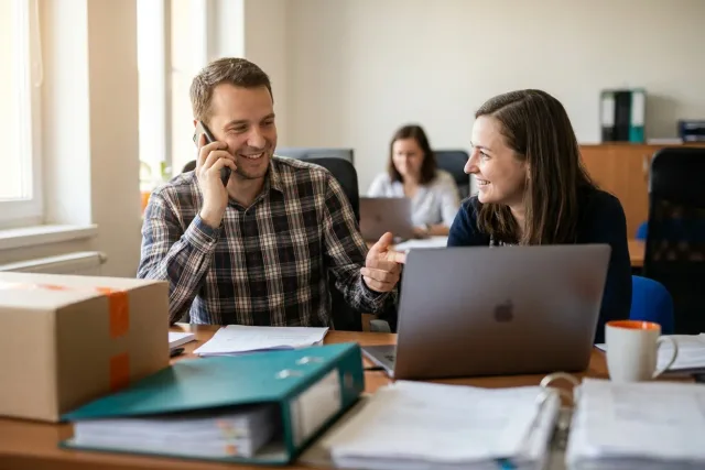 Obchodník telefonuje se zákazníkem a ukazuje kolegyni rozjednanou zakázku v notebooku v kanceláři.